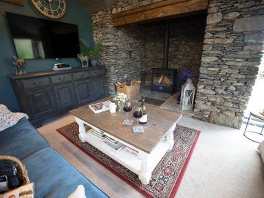 A living room with a wooden coffee table holding wine glasses and a bottle in front of a stone fireplace at The Owl's Shack in Kendal