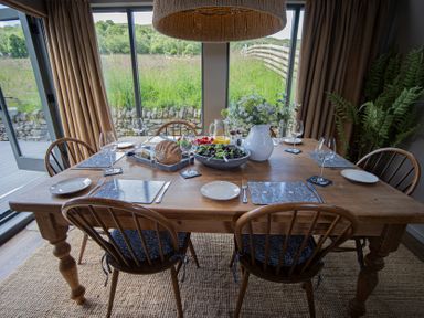 A wooden dining table set with plates forks knives wine glasses and food with large windows showing greenery at The Owl's Shack in Kendal