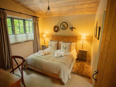 A bedroom with a wooden bed, bedside tables with lamps, a chair, and a window with curtains at The Owl's Shack in Kendal