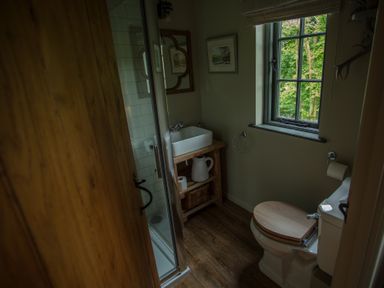 A bathroom with a wooden door wooden floor a toilet with wooden seat a sink on a wooden cabinet and a window showing green trees at The Owl's Shack in Kendal