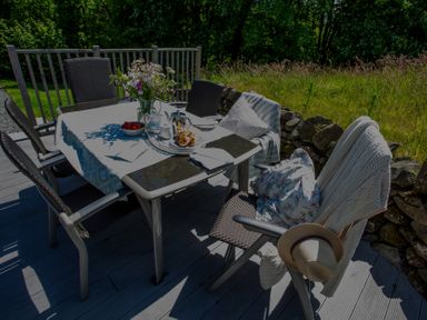 An outdoor dining table with chairs a vase of flowers and a tea set on a wooden deck with grass and stone wall outside at The Owl's Shack in Kendal