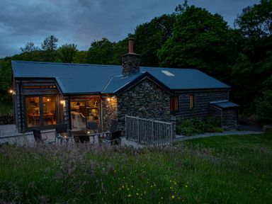 An exterior view of a stone and wood house with lit windows and outdoor seating surrounded by grass and trees at The Owl's Shack in Kendal