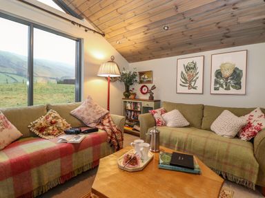 A living room with two sofas cushions and blankets a wooden coffee table with books and cups a floor lamp and a window showing a grassy landscape at The Badger's Shack in Garnett Bridge Longsleddale Near Kendal