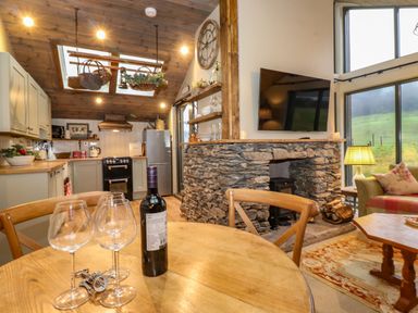 A kitchen and living area with a wooden dining table holding wine glasses and bottle near a stone fireplace with a mounted TV at The Badger's Shack in Garnett Bridge Longsleddale Near Kendal