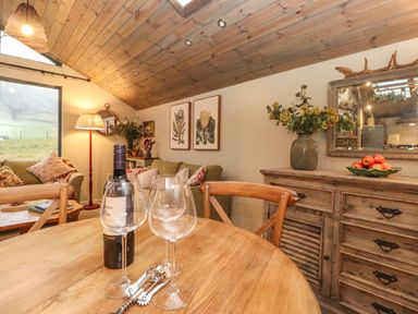 A dining table with wine glasses and a wine bottle in a living room with wooden ceiling and a sideboard with flowers and fruit at The Badger's Shack in Garnett Bridge Longsleddale Near Kendal