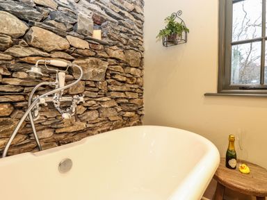 A bathroom with a white bathtub against a stone wall a wooden side table with a small bottle and glass and a window at The Badger's Shack in Garnett Bridge Longsleddale Near Kendal