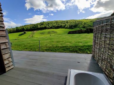 An outdoor bathtub on a wooden deck facing a green grassy hill under a partly cloudy sky at The Hare's Shack in Garnett Bridge Longsleddale Near Kendal