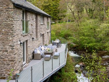 An outdoor deck with seating and dining area next to a stone house overlooking a stream with trees at The Old Water Mill in Kendal