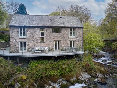 A stone house with a slate roof and a balcony overlooking a river and rocks at The Old Water Mill in Kendal