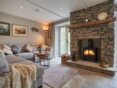 A living room with a stone fireplace and wood burning stove a grey sofa with pillows and a knitted throw a round wooden table with a bottle of wine and two glasses at The Old Water Mill in Kendal