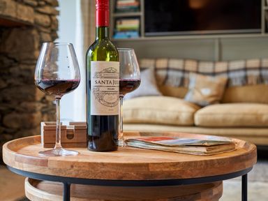 A wooden round table with two wine glasses filled with red wine a wine bottle and a folded magazine in a living room at The Old Water Mill in Kendal