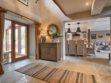 An entrance area with a wooden sideboard lamp and mirror next to a doorway leading to a dining area with wicker chairs and pendant lights at The Old Water Mill in Kendal