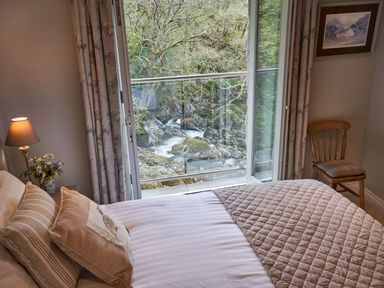 A bedroom with a bed pillows bedside lamp and chair next to an open window overlooking a rocky stream at The Old Water Mill in Kendal