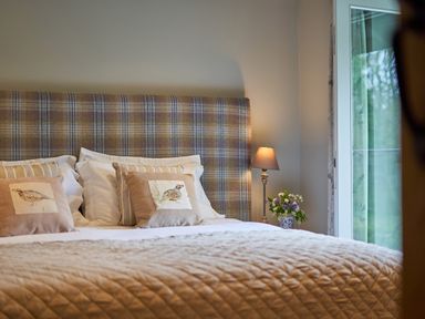 A bedroom with a plaid headboard two decorative bird pillows a bedside lamp and flowers by the window at The Old Water Mill in Kendal