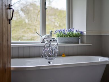 A white bathtub with a silver faucet and handheld shower with a yellow rubber duck and window with purple flowers on the sill at The Old Water Mill in Kendal