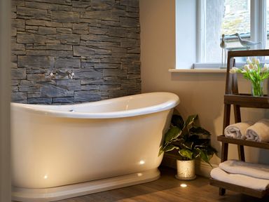 A bathroom with a white freestanding bathtub stone wall and a wooden shelf with towels and flowers at The Old Water Mill in Kendal