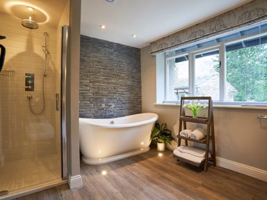 A bathroom with a freestanding bathtub against a stone wall and a glass enclosed shower next to a window with a wooden shelf holding towels and flowers at The Old Water Mill in Kendal