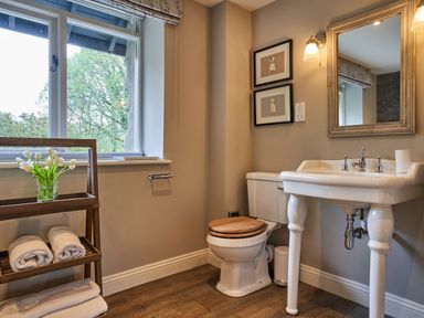 A bathroom with a toilet with a wooden seat a white sink a large mirror and a wooden shelf with towels and flowers at The Old Water Mill in Kendal