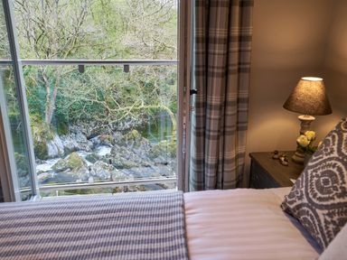 A bedroom with a bed pillow lamp and flowers on a nightstand overlooking a river and rocks at The Old Water Mill in Kendal