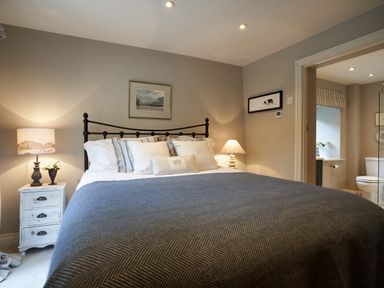 A bedroom with a bed with pillows and a grey blanket two bedside tables with lamps a painting above the headboard and an adjoining bathroom at The Old Water Mill in Kendal