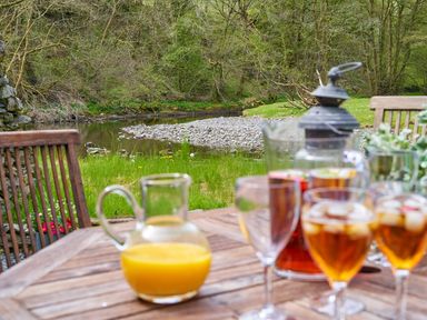 An outdoor wooden table with glasses of iced tea and a jug of orange juice near a river and trees at The Old Water Mill in Kendal