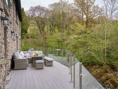 An outdoor deck with wicker furniture and a glass railing next to a stone wall overlooking trees at The Old Water Mill in Kendal