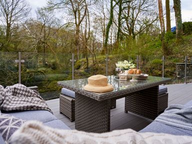 An outdoor seating area with a glass table a hat flowers and baked goods surrounded by trees at The Old Water Mill in Kendal