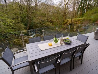 An outdoor dining table with chairs on a deck overlooking trees and a rocky stream at The Old Water Mill in Kendal