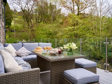 An outdoor seating area with cushioned chairs and a glass table set with scones tea and flowers surrounded by trees at The Old Water Mill in Kendal
