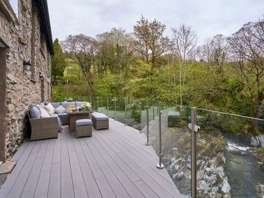 An outdoor patio with wicker seating and a glass railing overlooking a forest and rocky stream at The Old Water Mill in Kendal