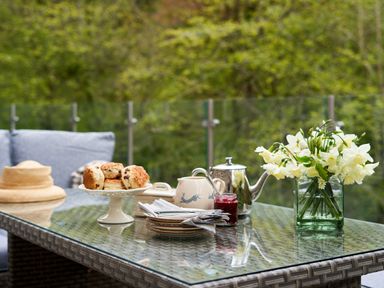 An outdoor glass table with scones on a cake stand a teapot a jar of jam a milk jug plates with cutlery and a vase of white flowers at The Old Water Mill in Kendal