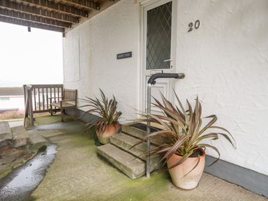 A covered entrance with steps two potted plants a wooden bench and a door labeled sandy toes at Rhosneigr