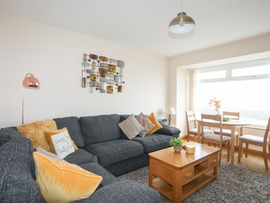 A living room with a dark sofa and cushions a wooden coffee table and a dining table with chairs near a window at Sandy Toes in Rhosneigr