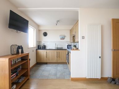 A kitchen with wooden cabinets a tiled floor a clock and a microwave near a wooden shelf with books and a television mounted on the wall at Sandy Toes in Rhosneigr