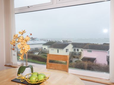A dining table with a yellow flower pot and a fruit basket with green apples by a window overlooking houses and the sea at Sandy Toes in Rhosneigr