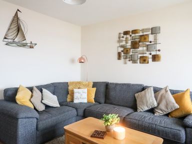 A living room with a dark gray corner sofa with yellow and striped cushions wooden coffee table with a plant and candle metal sailboat wall decoration and metal abstract wall art at Sandy Toes in Rhosneigr