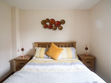 A bedroom with a wooden bed a yellow and gray striped bedspread two wooden bedside tables with lamps and wall decor above the bed at Sandy Toes in Rhosneigr