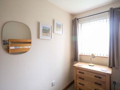 A bedroom corner with a wooden dresser under a window with curtains a lamp on the dresser a wall mirror and two framed pictures on the wall at Sandy Toes in Rhosneigr