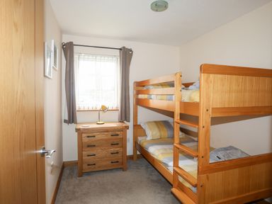A bedroom with wooden bunk beds a wooden chest of drawers a window with curtains and a desk lamp at Sandy Toes in Rhosneigr