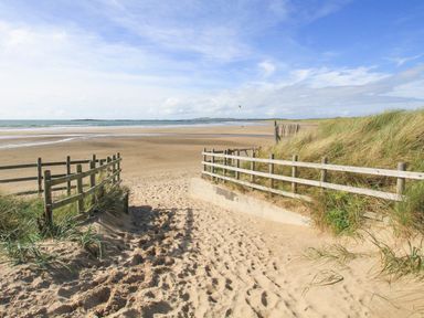 A sandy path with wooden fences leading to a beach with sea and grassy dunes in the background at Sandy Toes in Rhosneigr