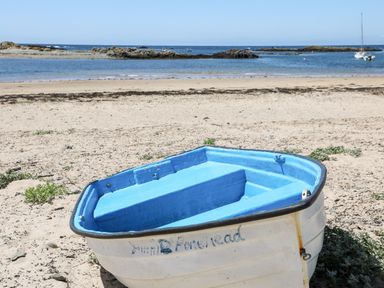 A small blue and white boat on a sandy beach with rocks and boats in the water in the background at Sandy Toes in Rhosneigr