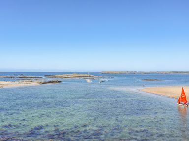 A coastal scene with clear water sandbanks and small boats including a red sailboat near the shore at Sandy Toes in Rhosneigr