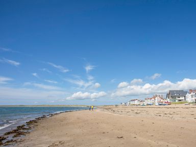 A sandy beach with two people walking near the water and houses along the shore at Sandy Toes in Rhosneigr