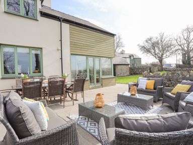 An outdoor patio with wicker seating and glass tables next to a house with green window frames at Dolwaenydd in Brynsiencyn