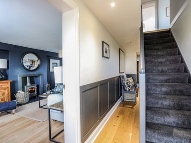 A hallway with wooden floor a chair and framed pictures next to carpeted stairs and a living room with fireplace at Dolwaenydd in Brynsiencyn