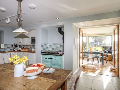 A kitchen with wooden table and chairs flowers on table and an open doorway leading to a dining area with blue chairs and a TV at Dolwaenydd in Brynsiencyn