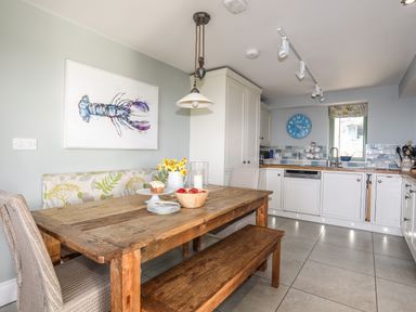 A kitchen with white cabinets a wooden table with chairs and bench a blue clock and a lobster painting on the wall at Dolwaenydd in Brynsiencyn