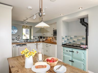 A kitchen with a wooden table holding flowers a candle cake and apples a blue stove wooden counters and cream cabinets at Dolwaenydd Brynsiencyn