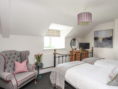 A bedroom with two single beds a gray armchair a side table with flowers a wooden dresser a round mirror and a painting at Dolwaenydd in Brynsiencyn