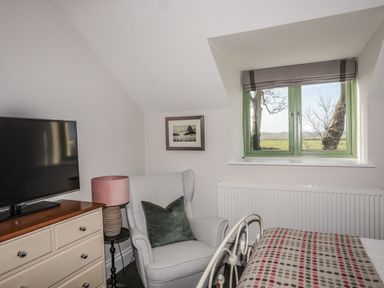 A bedroom with a television on a dresser an armchair with a pillow a lamp a window and a bed with a patterned blanket at Dolwaenydd in Brynsiencyn